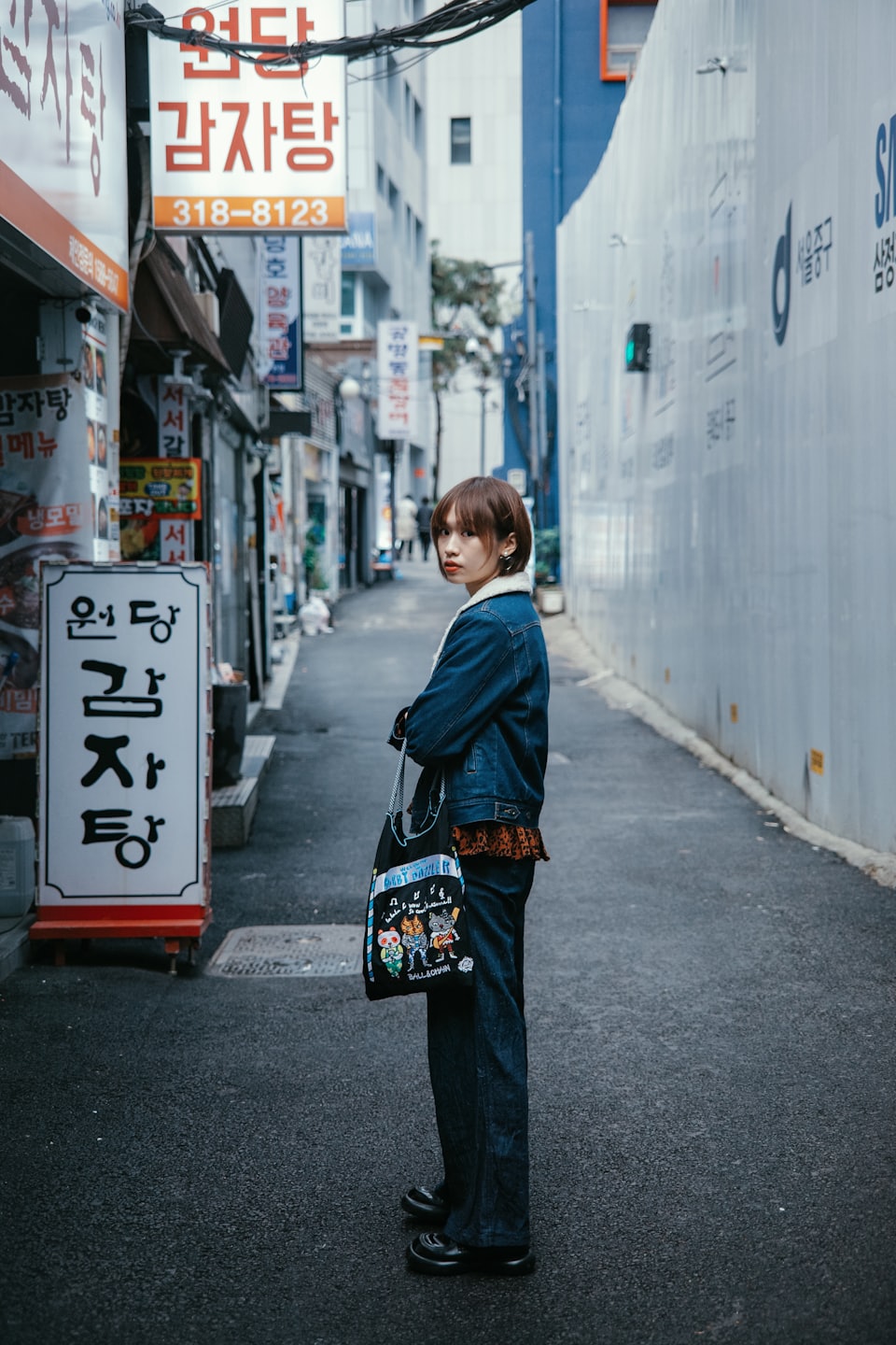 Woman in denim jacket with cartoon tote bag in Korean alleyway.