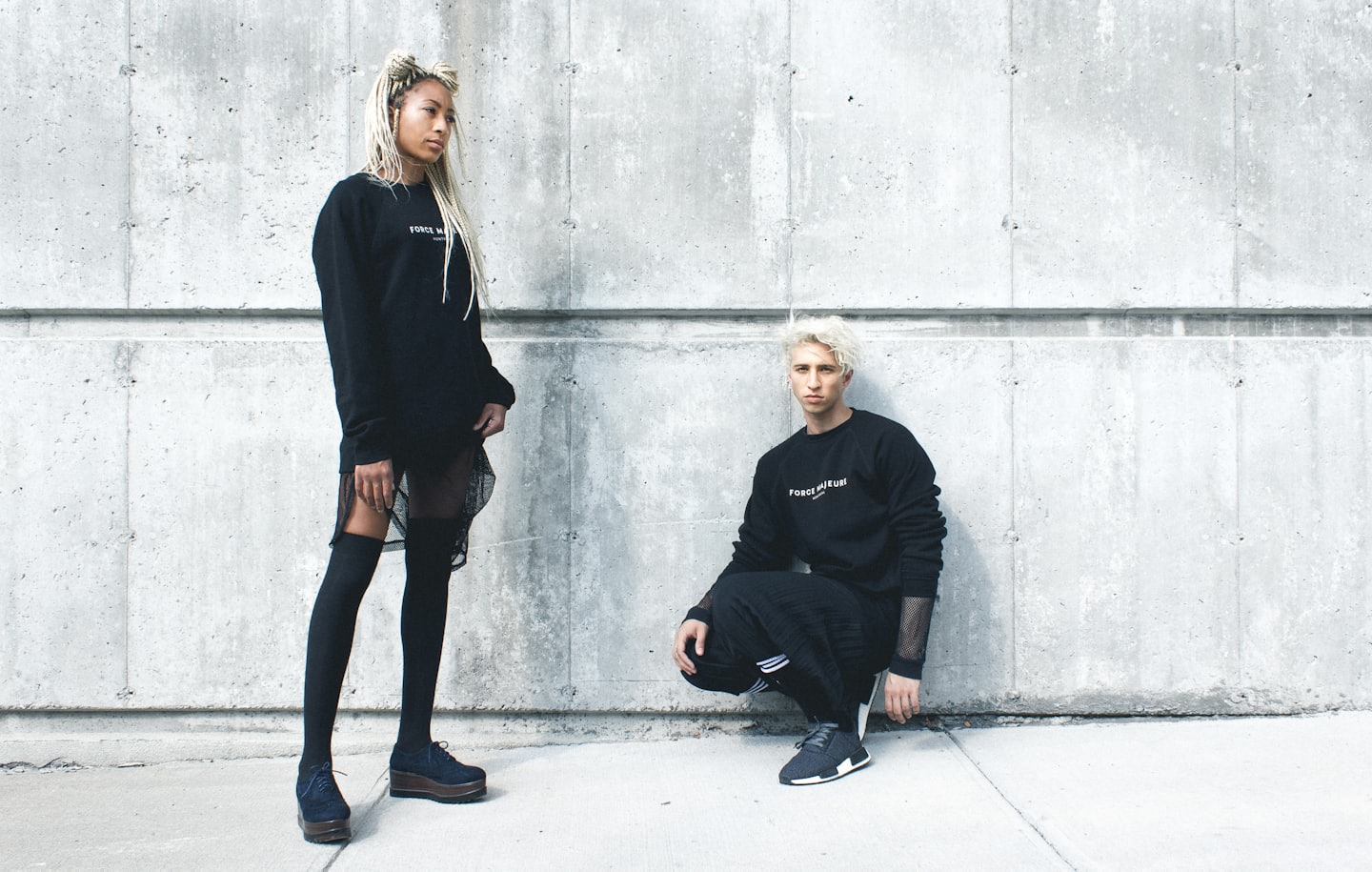 Woman and man in streetwear posing by concrete wall.